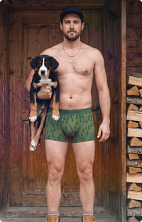 Man holding a puppy wearing green patterned underwear in a rustic setting